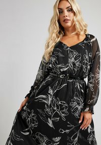 Zwarte bloemenjurk met doorschijnende lange mouwen, met een cinched taille met een riem. Witte bloemenpatroon en textuurdetail.