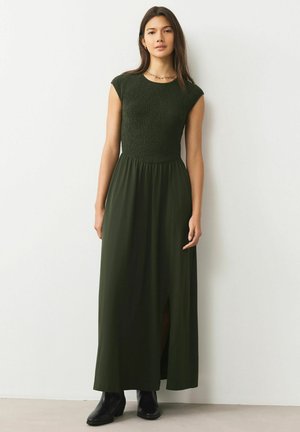 Dunkelgrünes Maxikleid mit gerafftem Oberteil und kurzen Ärmeln. Mit einem Frontschlitz, der bis zu den Knöcheln reicht, kombiniert mit schwarzen Stiefeln bis zum Knöchel.
