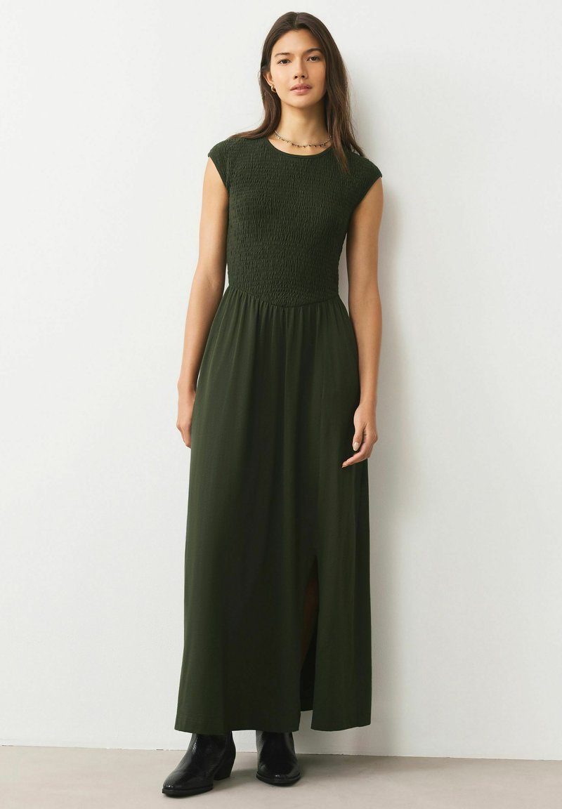 Donkergroene maxi-jurk met een geplooide bustier en korte mouwen. Heeft een split aan de voorkant en valt tot aan de enkels, gecombineerd met zwarte enkellaarzen.