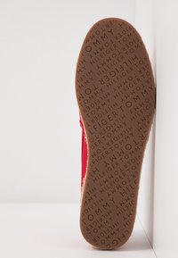 Semelle en caoutchouc avec logo embossé, tige en tissu rouge et coutures sur le côté. Design plat adapté à un usage décontracté.