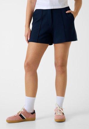 Person trägt marineblaue Shorts, weiße Socken, pinke Turnschuhe und ein weißes Oberteil, steht mit einer Hand in der Tasche vor einem schlichten Hintergrund.