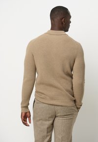Beige gerippter Pullover mit Kragen, lange Ärmel und taillierter Passform, kombiniert mit strukturierten hellbraunen Hosen.