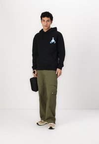 Black hoodie with blue graphic logo, relaxed fit, paired with olive cargo pants featuring pockets, and beige sneakers. Holding a black pouch.