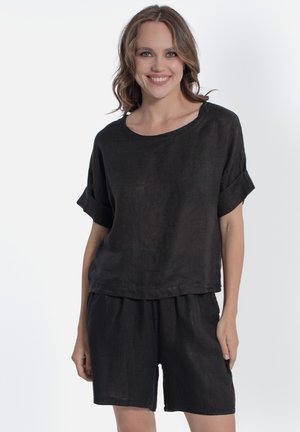 Donna sorridente che indossa una maglia nera larga a maniche corte con polsini risvoltati e shorts neri abbinati su uno sfondo bianco uniforme.