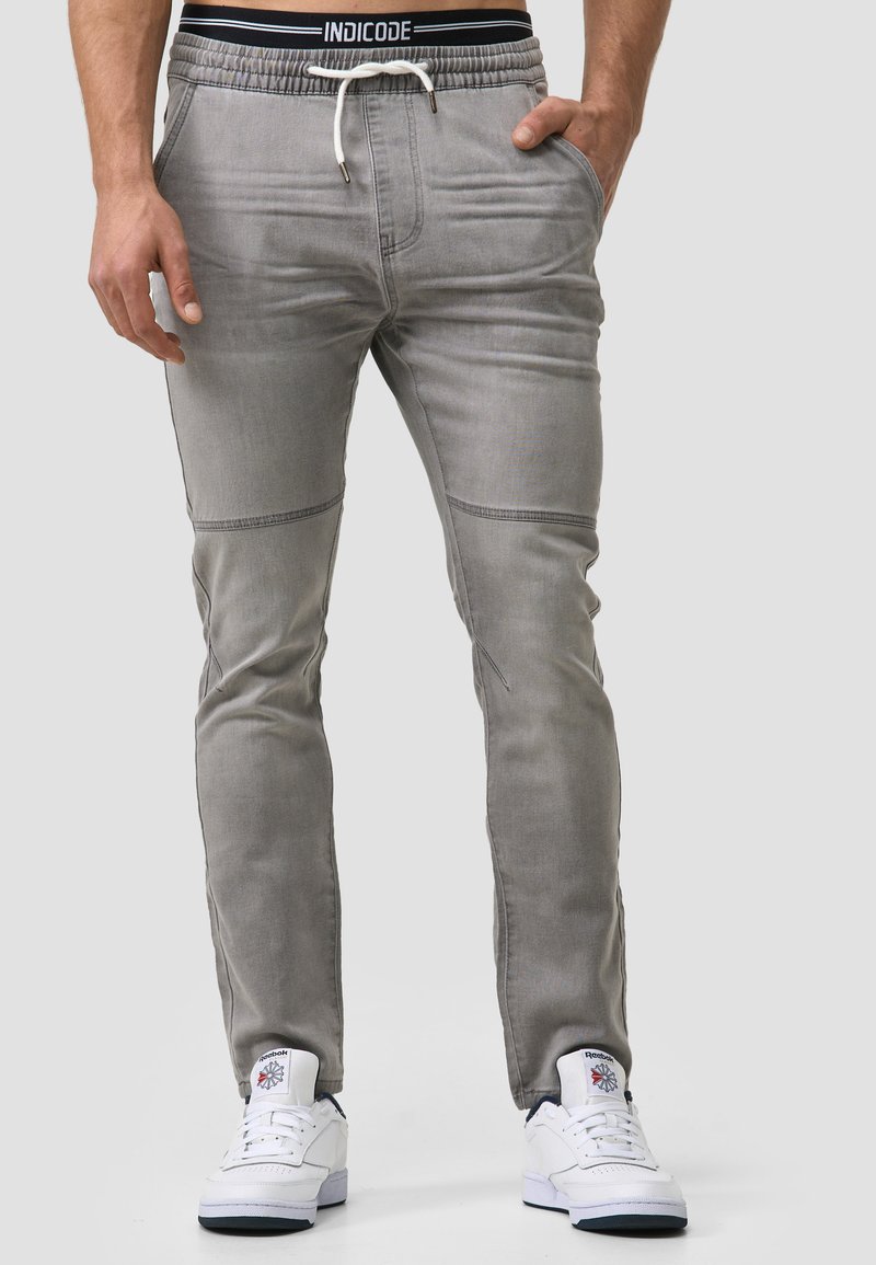Pantalones jogger grises con cintura elástica y cordón, que cuentan con bolsillos laterales y costuras en contraste, combinados con zapatillas blancas.