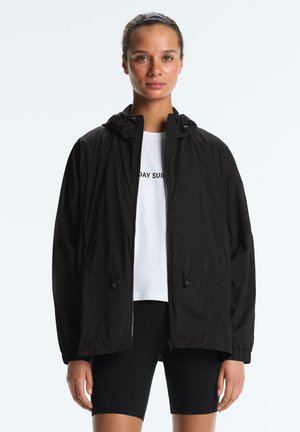 Kobieta ubrana w czarną bluzę z kapturem na zamek, na białej koszulce i czarnych spodenkach rowerowych, stojąca na jasnym, jednolitym tle.