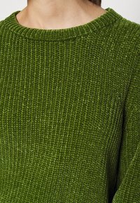 Gros plan d'une personne portant un pull en tricot vert texturé avec un col rond, montrant le dos et la partie supérieure de l'épaule.