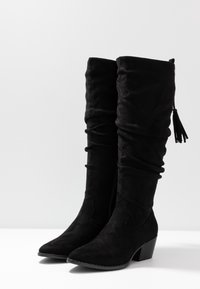Bottes hautes jusqu'au genou en daim noir avec un design effondré, un bout pointu et un petit talon bloc, ornées d'un détail à pampille sur la tige.