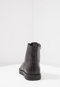 Stivale alla caviglia in pelle nera con zip sul retro, maniglia per l'estrazione e suola spessa, mostrato da dietro su una superficie bianca e sfondo bianco.