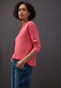 Pull en maille rose corail à manches longues avec poignets côtelés, associé à un jean en denim bleu avec deux poches avant et un détail bouton.