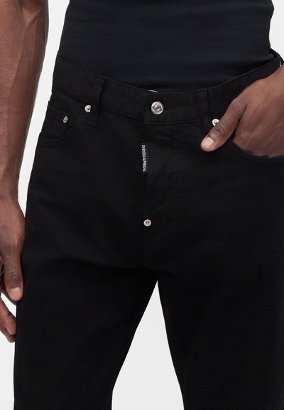 Homme portant un jean noir avec des boutons argentés et une chemise noire, une main dans la poche et l'autre détendue à côté.