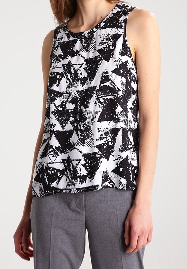 Femme portant un haut sans manches à motif abstrait noir et blanc et un pantalon gris, debout devant un fond clair uni.