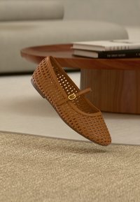 Καφέ πλεκτό παπούτσι Mary Jane με χρυσή πόρπη σε λουράκι, αιωρούμενο πάνω από μπεζ υφασμάτινο χαλί, ξύλινο τραπεζάκι καφέ και φόντο καναπέ.
