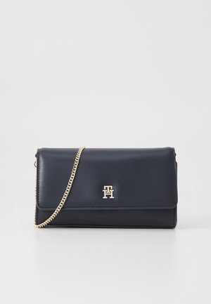 Pochette en cuir noir avec bandoulière chaîne dorée et logo "TH" doré centré sur le rabat avant, sur fond uni.