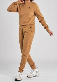 Felpa in cotone marrone e pantaloni jogger abbinati caratterizzati da un logo discreto. L'outfit ha una vestibilità comoda, polsini a coste e una texture morbida. Le scarpe sono bianche con dettagli grigi.