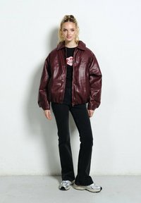 Burgundowa kurtka z sztucznej skóry z dużym kołnierzem, zapinana na zamek z przodu, w luźnym kroju, zestawiona z czarną bluzką i rozszerzanymi czarnymi dżinsami.