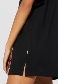 Vue de profil gauche d'une personne portant une robe noire avec une petite fente latérale et une petite étiquette blanche près de l'ourlet.