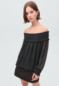 Svart off-shoulder-blus med smockad midja och långa genomskinliga ärmar. Stylingen inkluderar en svart jeanskirt med sidogång.