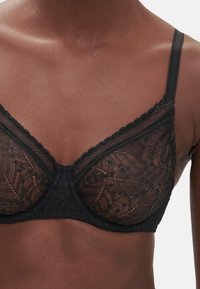 Svart spetsbehå med genomskinliga kupor, prydd med blommönster och vågig kant. Justerbara axelband och stöd med underwire.