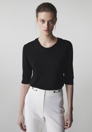 Jeune femme aux cheveux attachés, portant un haut noir à manches courtes et un pantalon blanc taille haute, sur un fond uni.