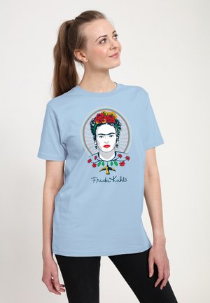 Ung kvinde iført lyseblå T-shirt med farverig Frida Kahlo-portræt og blomster, stående mod en ensfarvet baggrund og kiggende op.