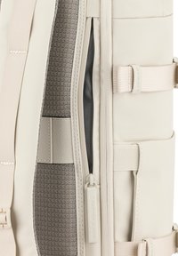 Interiér bílé batohu s texturovanou šedou podšívkou, černou kapsou se zipem a nastavitelné béžové popruhy. Kroužek na zipu má logo značky.