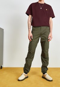Ciemnoczerwony T-shirt, luźny krój. Oliwkowe spodnie cargo z bocznymi kieszeniami. Beżowe buty wsuwane z ciemną podeszwą i brązowymi akcentami.