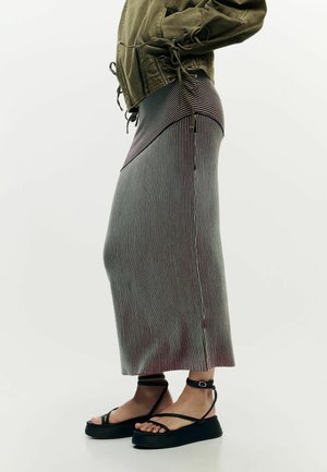 Isik, kes kannab oliivrohelise jaku, kõrge vöökohaga triibulist midi seelikut ja musti platvormsandaale pahkluu rihmadega.