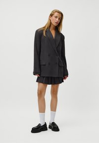 Blazer duplo em cinza escuro combinado com uma saia plissada, acentuado por mocassins pretos e meias brancas, em um fundo neutro.