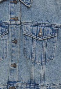 Ljusblå denimjacka med detalj av framsidan, metallknappar och fack för bröstfickor, synlig sömnad och sliten textur.