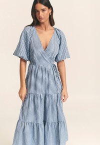 Sinivalkoinen ruutukuosinen mekko, jossa syvä V-pääntie, lyhyet liehukkeet ja kerroksittainen hameen muotoilu. Vyötäröllä solmittava yksityiskohta.