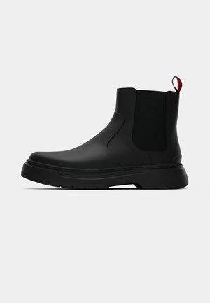 Zwarte enkelboot met een gladde afwerking, elastische zijpanelen en een contrasterende rode treklus, met een dikke rubberen zool en een afgeronde teen.