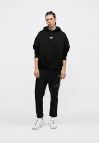 Felpa nera in cotone con tasca frontale e logo, abbinata a pantaloni jogger neri con un dettaglio laterale testurizzato e scarpe da ginnastica bianche.