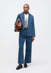 Persona che indossa una giacca di denim e jeans a gamba larga con una camicia a righe, mocassini neri e tiene in mano una grande borsa di pelle marrone.