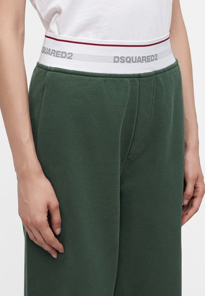 Personne portant un pantalon de survêtement vert foncé avec une ceinture élastique blanche siglée DSQUARED2, associé à une chemise blanche rentrée.