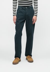 Jeans de algodão azul escuro com um design de perna reta, cinco bolsos e ferragens em metal, combinados com sapatos de camurça castanhos. Camisola de manga longa às riscas visível.