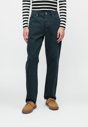 Granatowe dżinsy bawełniane o prostym kroju, pięć kieszeni i metalowe okucia, w zestawieniu z brązowymi zamszowymi butami. Widoczna jest też koszulka w paski z długim rękawem.