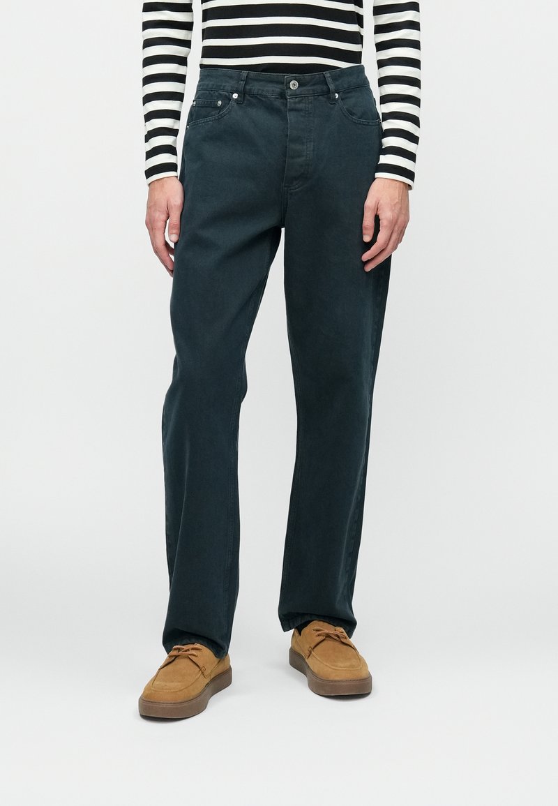 Jeans de algodão azul escuro com um design de perna reta, cinco bolsos e ferragens em metal, combinados com sapatos de camurça castanhos. Camisola de manga longa às riscas visível.