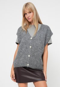 Gray knitted vest with a v-neck, short sleeves, and four white buttons. Paired with a gray t-shirt and a fitted burgundy faux leather skirt.