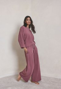 Mujer de pie descalza con una blusa suelta de manga larga color malva y pantalones de pierna ancha frente a una pared lisa y clara y suelo alfombrado.