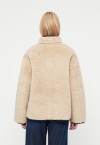 Veste en fausse fourrure beige avec une finition douce et texturée, grand col, épaules tombantes et coupe décontractée. Coutures apparentes le long des bords.