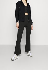 Sudadera negra con cremallera y diseño cropped, combinada con pantalones negros de campana. Zapatillas blancas con acentos plateados y amarillos completan el look.