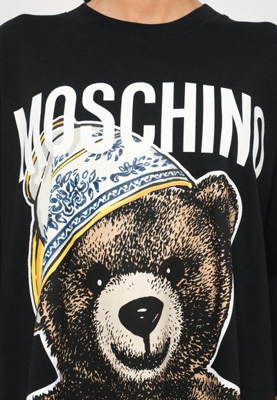 Melna T-krekls ar karikatūras lāča grafiku un zilu un dzeltenu rakstainu cepuri. Logo "MOSCHINO" ir izteikti redzams virs lāča.