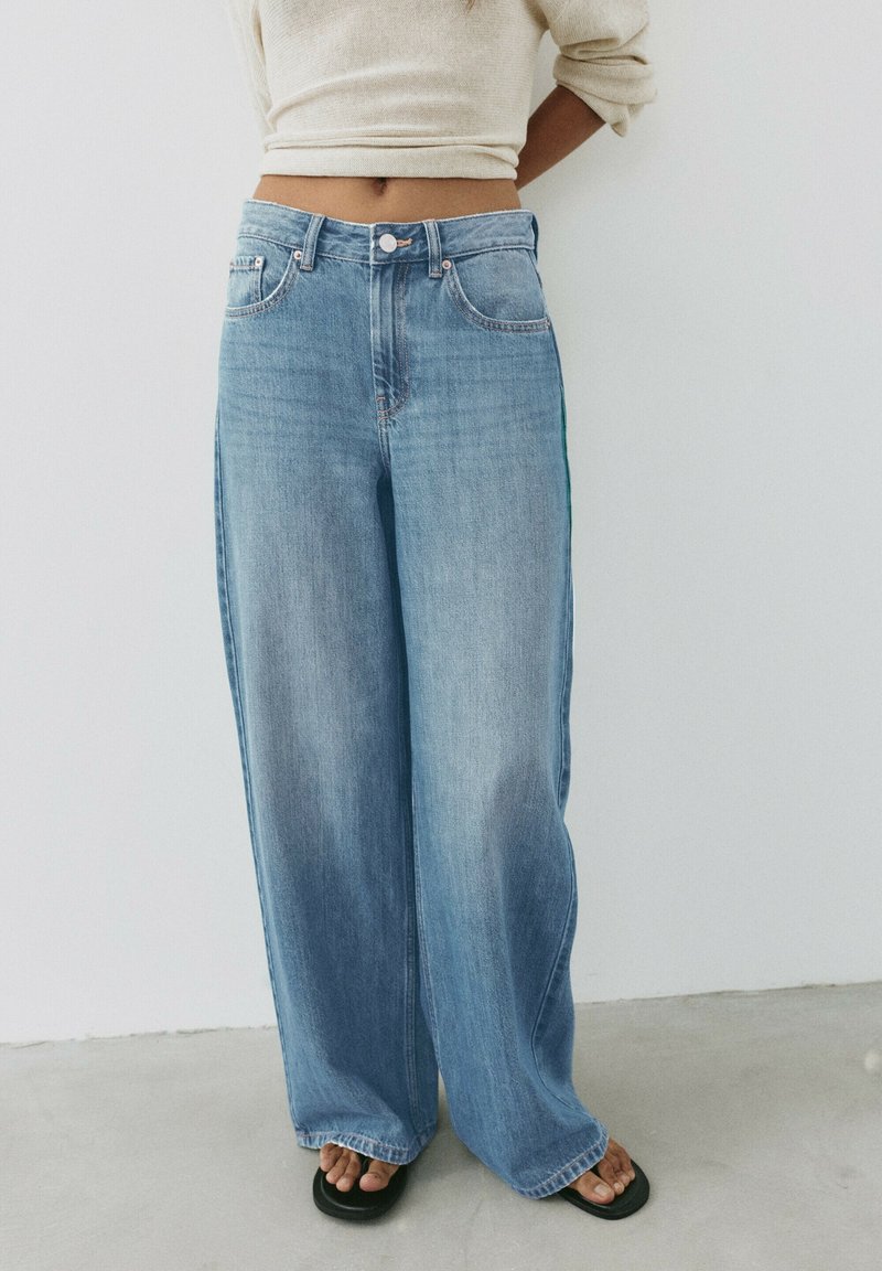 Femme portant un jean large bleu clair, un pull court crème et des sandales noires, debout sur un sol gris contre un mur blanc.