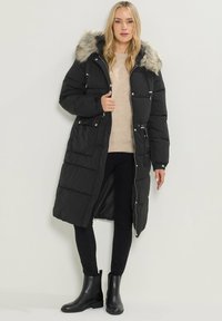 Abrigo de plumas largo y negro con capucha de piel desmontable, cierre de cremallera, bolsillos delanteros y diseño acolchado. Combinado con un suéter beige y pantalones negros.