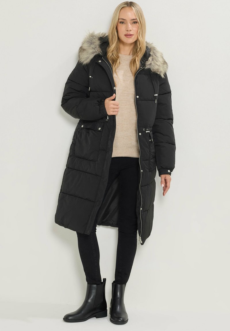 Abrigo de plumas largo y negro con capucha de piel desmontable, cierre de cremallera, bolsillos delanteros y diseño acolchado. Combinado con un suéter beige y pantalones negros.