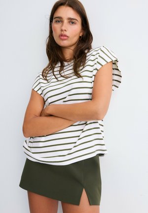 Jeune femme aux longs cheveux bruns, portant un haut rayé blanc et vert et une mini-jupe vert foncé avec une fente à l'avant, posant bras croisés.