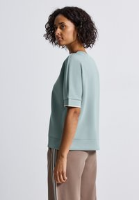 Sweatshirt bleu clair à manches courtes avec un col rond, texture lisse et poignets côtelés, associé à un pantalon beige avec des rayures latérales.