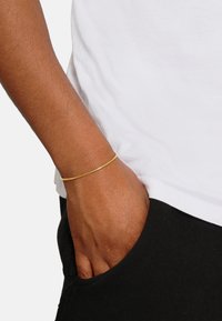 Goldener Armreif an einem Handgelenk mit gleichmäßiger Textur. Die Hand ist entspannt und ruht in einer Tasche, neben einem weißen Hemd und einer schwarzen Hose.