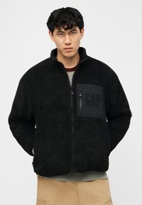 Νεαρός άνδρας φορώντας μαύρο μπουφάν από φλις με φερμουάρ στην τσέπη στο στήθος, μπεζ παντελόνι και τα χέρια στις πλαϊνές τσέπες, κοιτάζοντας προς τα εμπρός.